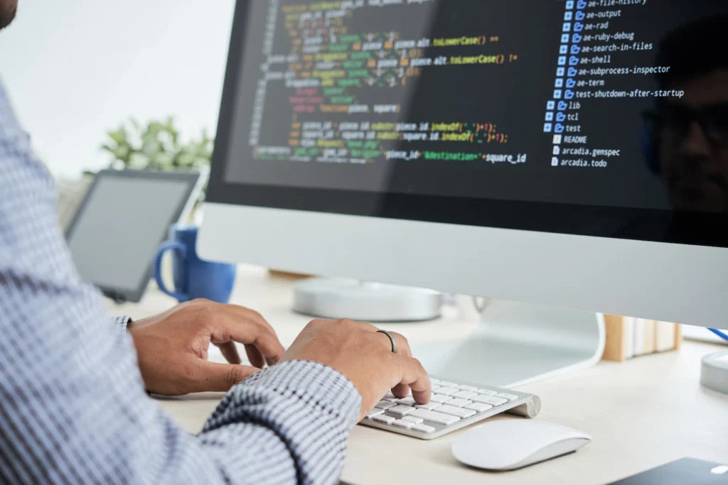 Programmierer tippt Code auf der Tastatur vor einem Monitor mit Quellcode einer Website oder Software.
